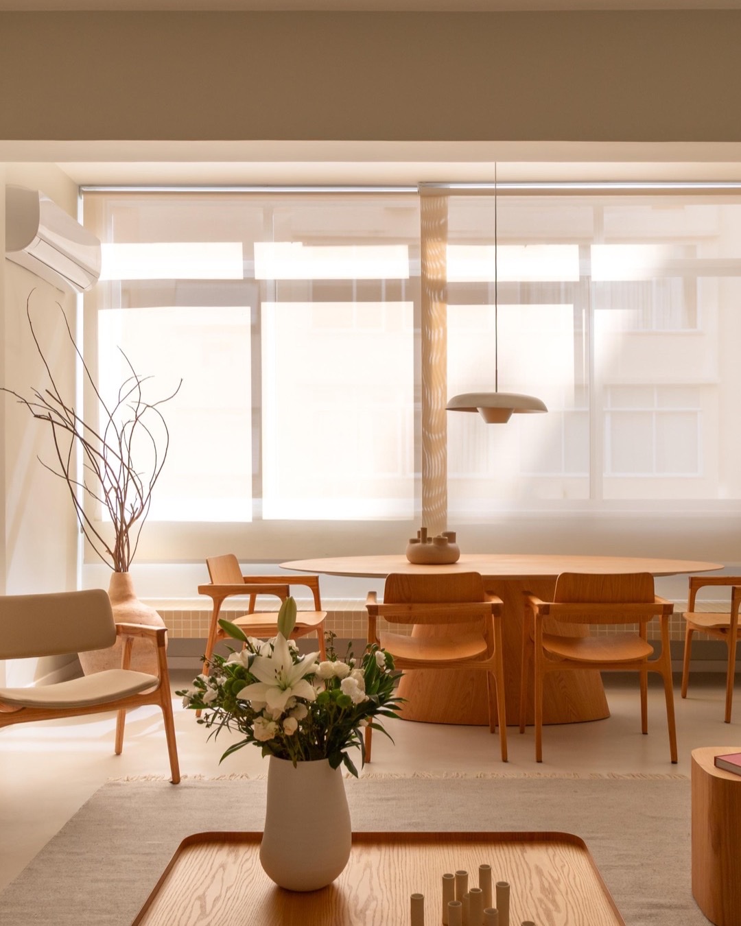 Sala de jantar com mesa de madeira, cadeiras claras, pendente e grande janela com luz natural