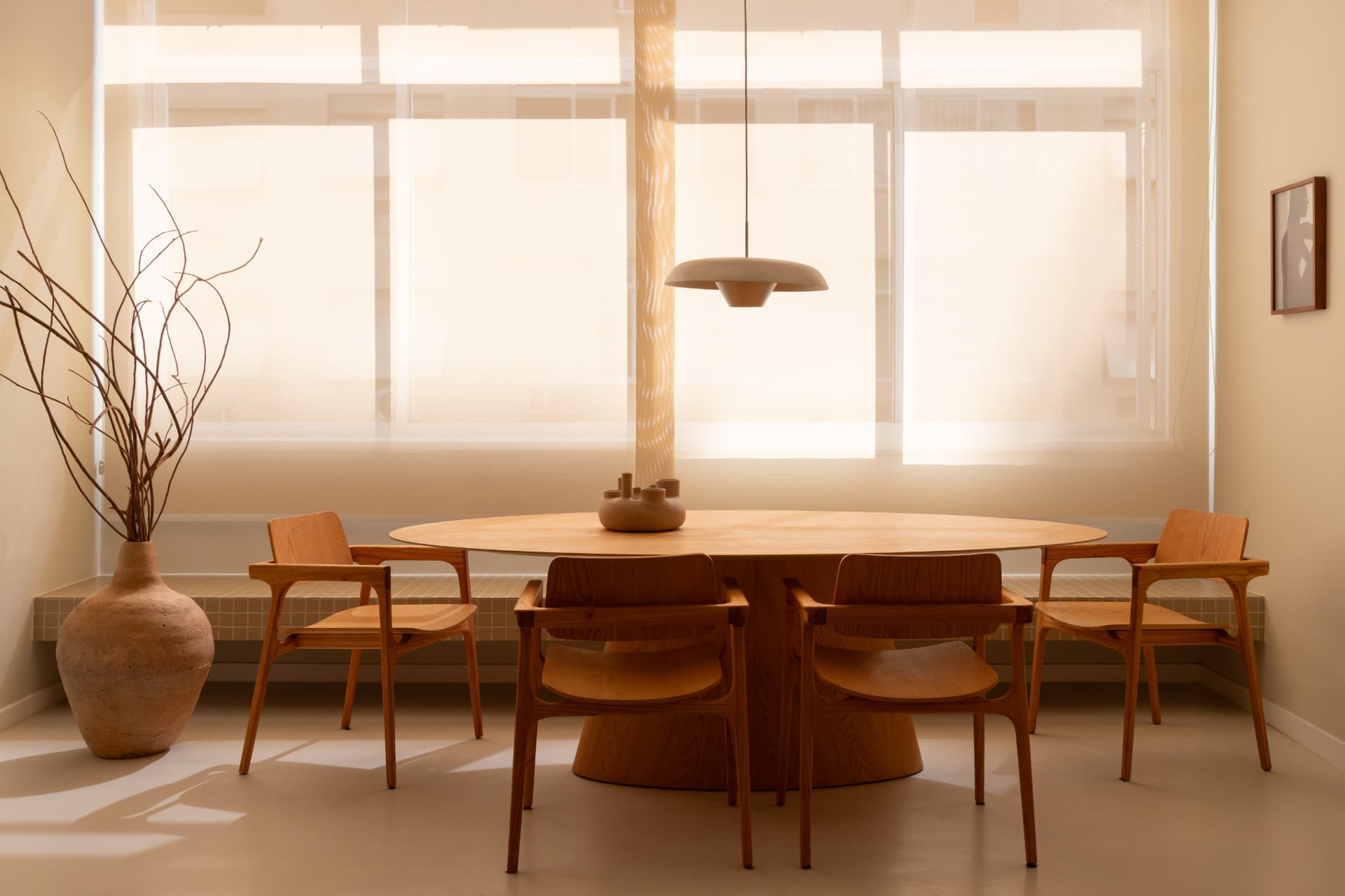 Sala de jantar com mesa oval de madeira, cadeiras de braço, pendente central e luz natural difusa pela janela