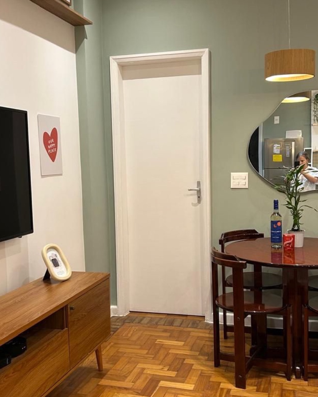 Sala integrada à copa com parede verde, espelho orgânico, pendente de madeira e piso em taco