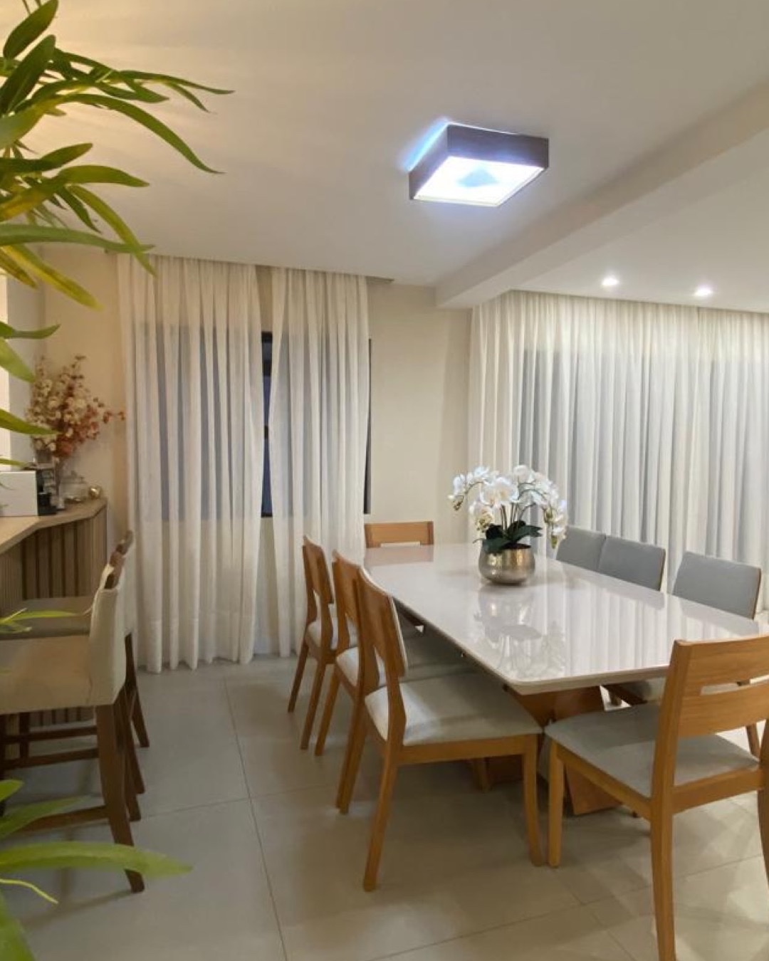 Sala de jantar clara com mesa ampla, cadeiras de madeira, cortinas e luminária quadrada