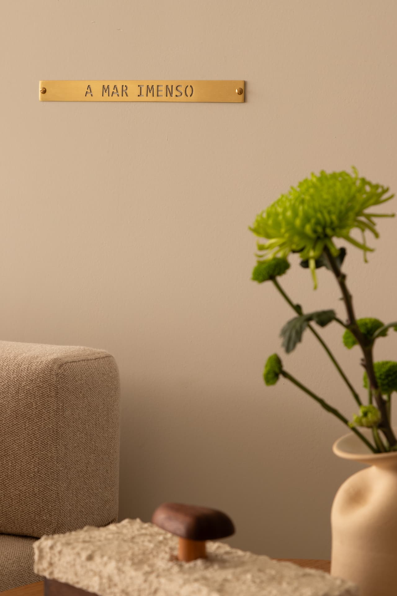 Detalhe de parede em tom areia com placa de latão, vaso de cerâmica com flores verdes e mesa lateral texturizada