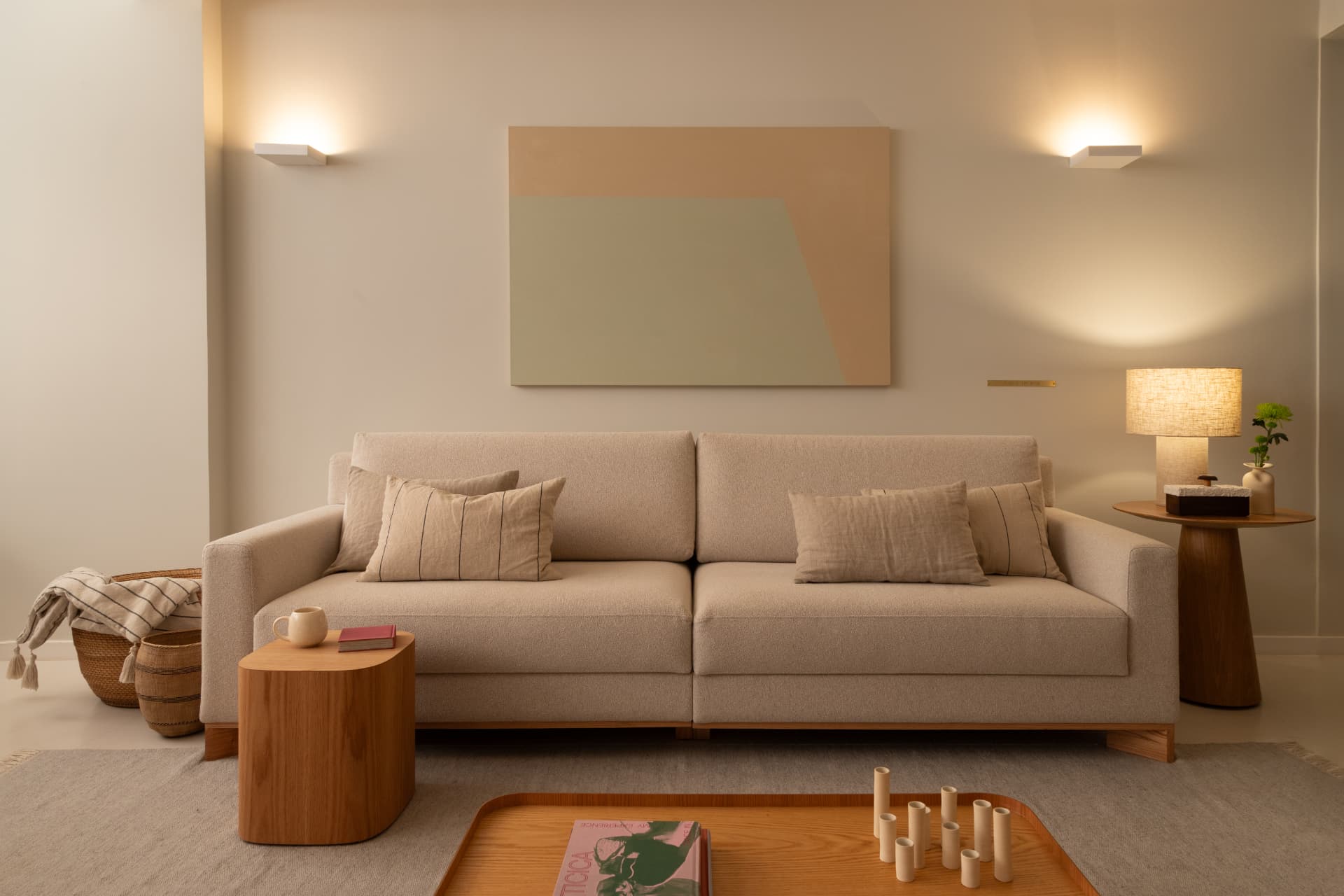 Sala de estar com sofá claro, arandelas de parede, quadro abstrato, mesas laterais de madeira e tapete neutro