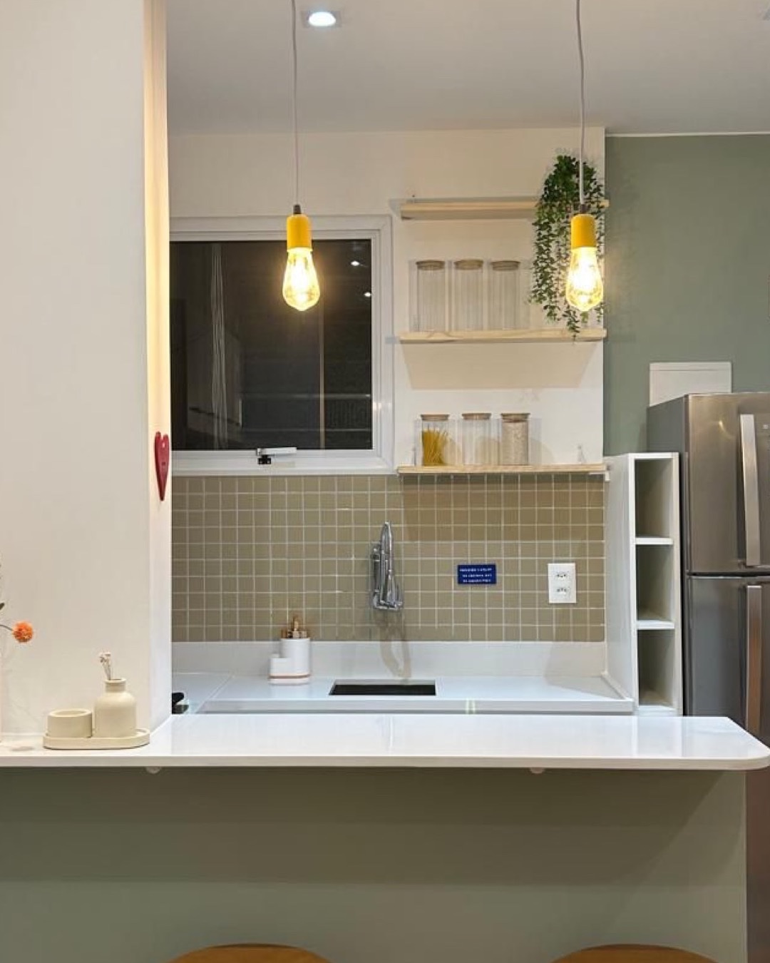 Cozinha com bancada bar branca, pendentes amarelos, prateleiras de madeira e azulejo verde-oliva